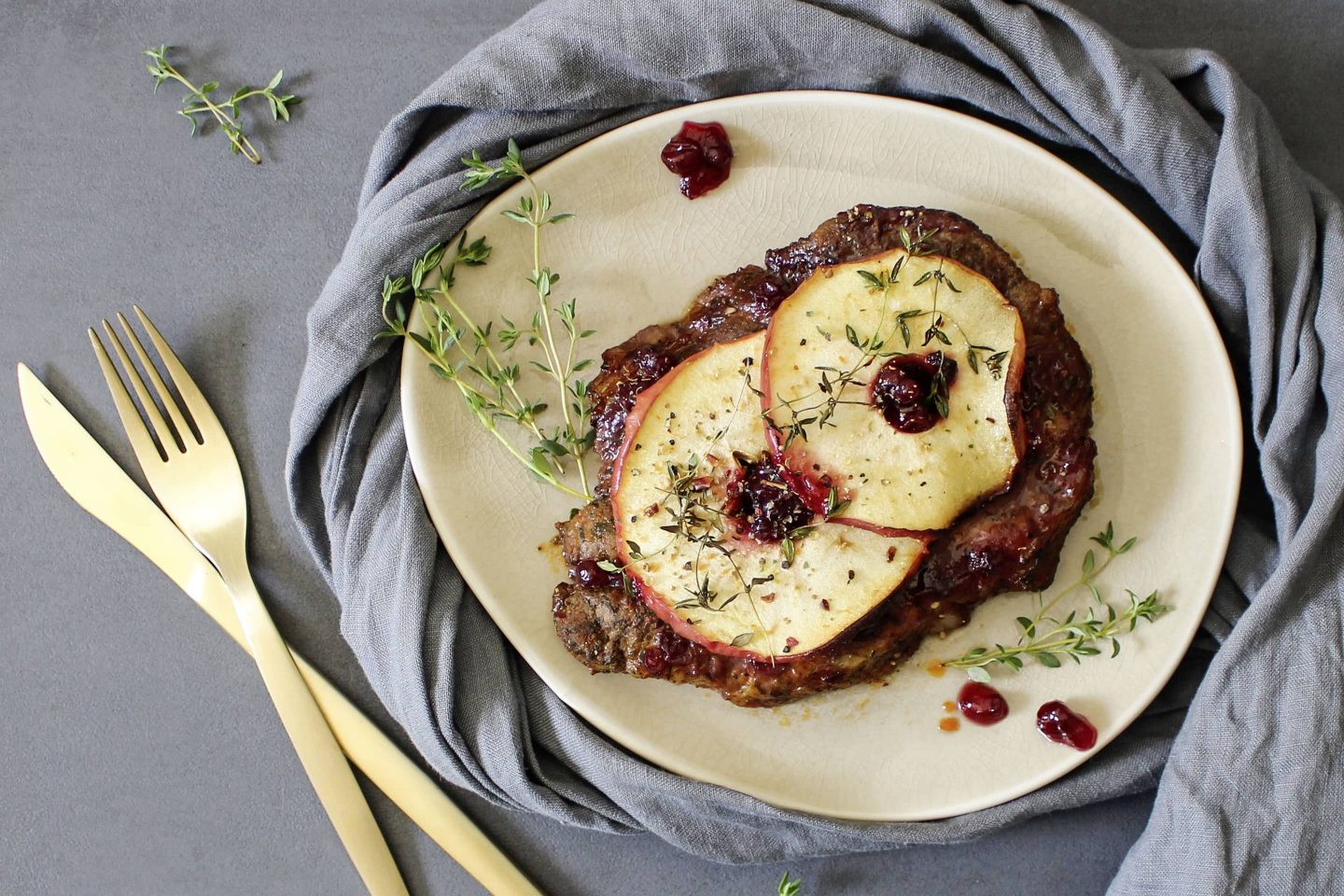 Chop with apple slices and cranberries garnished on a light plate with a gray napkin and a golden bowl