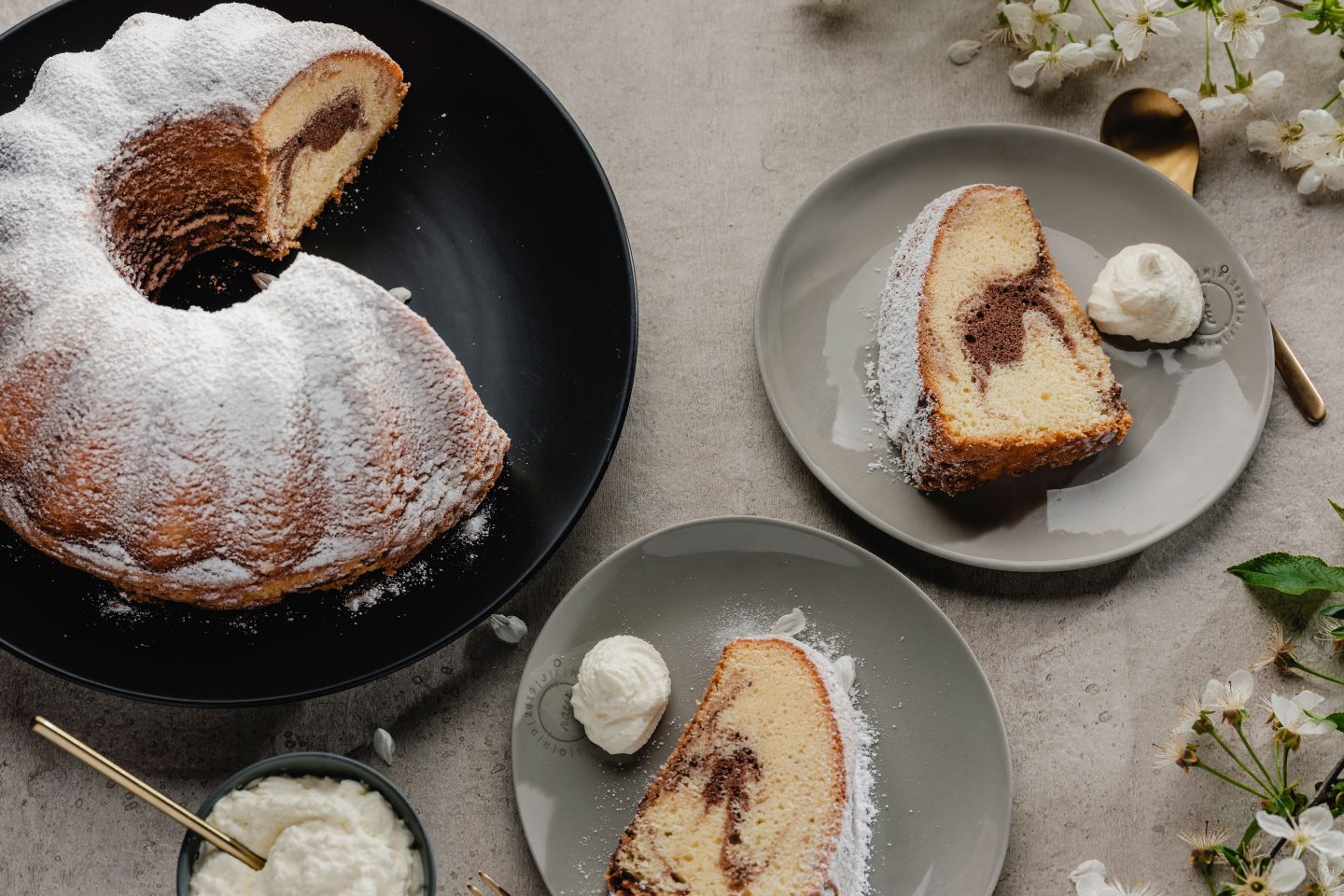 Marble Guglhupf on a black plate and 2 pieces of cake on small plates with whipped cream, golden forks and apple blossoms