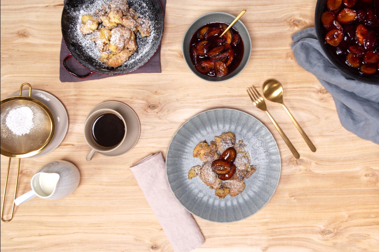 Kaiserschmarrn on a blue plate sprinkled with icing sugar, with pepper plums in a blue bowl and pan