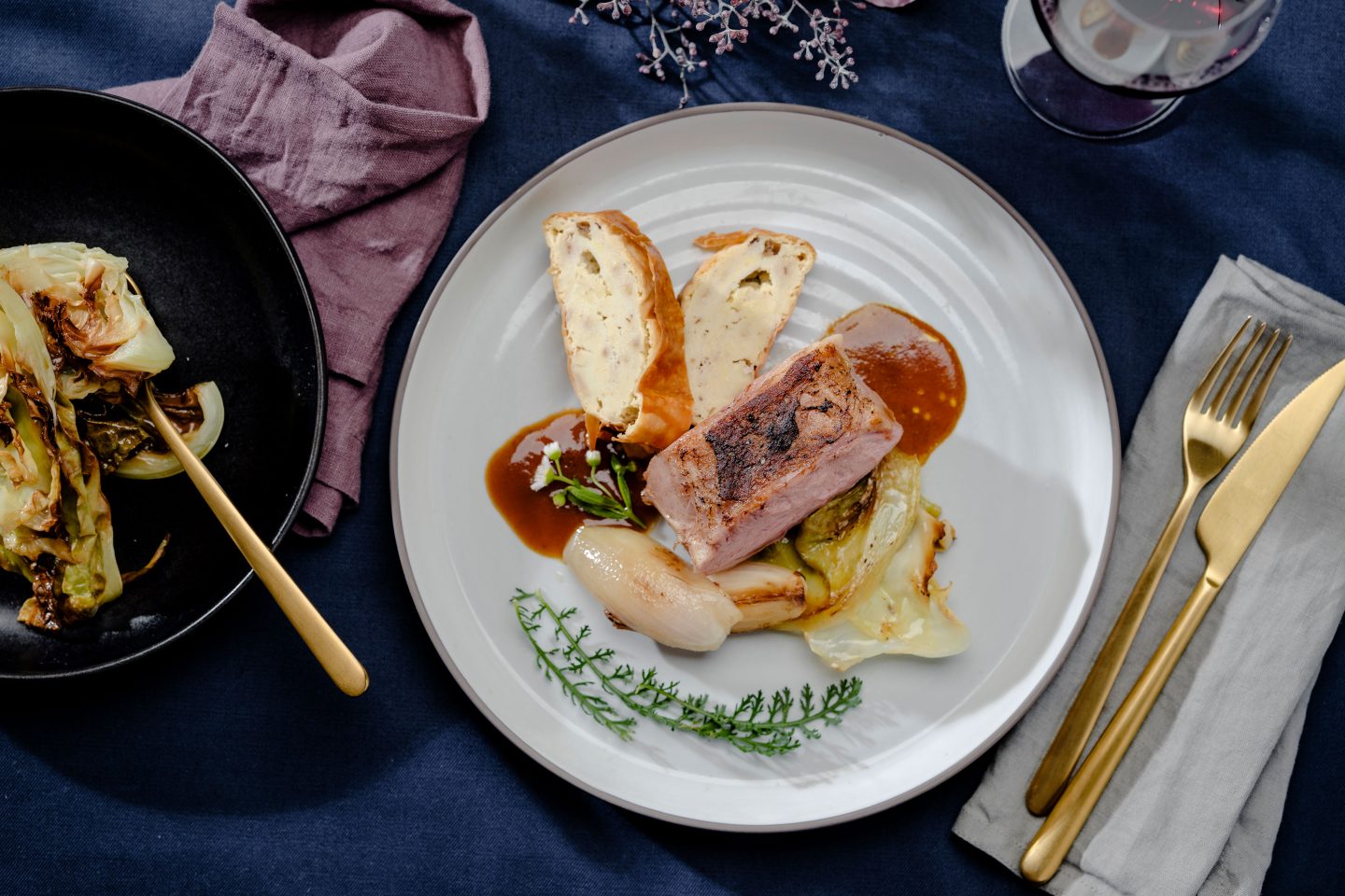 Roasted veal on roasted cabbage with shallots and semolina strudel on a beige plate with a glass of red wine and next to it a black plate with cabbage from the oven