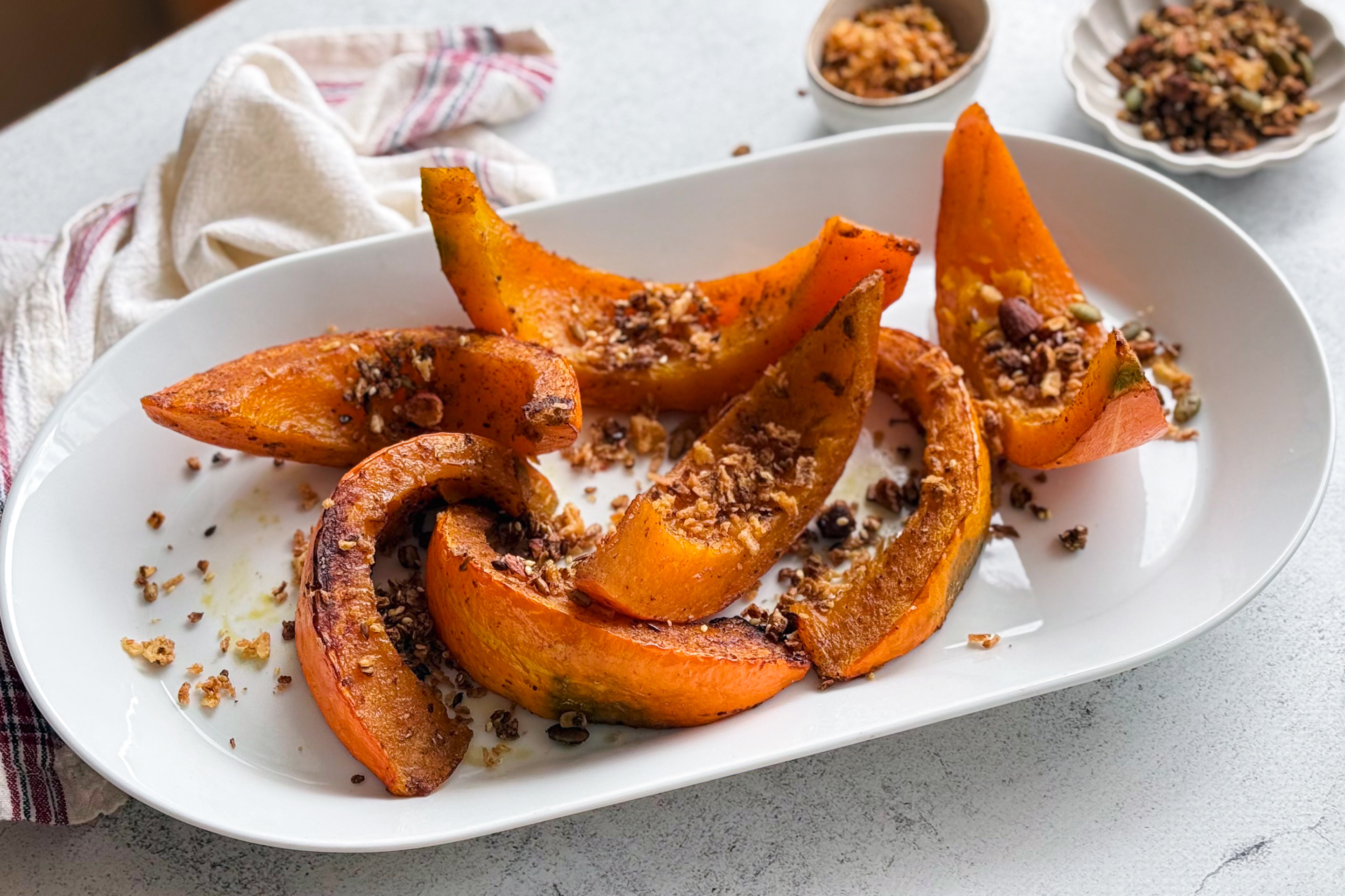 Roasted pumpkin wedges garnished with granola, nuts, and seeds, served with a drizzle of olive oil.